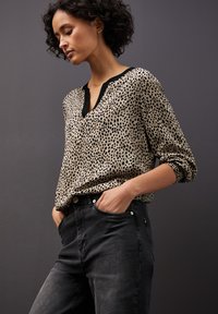 Bluzka w zwierzęcy wzór w beżowym kolorze z czarnymi plamami, z dekoltem w serek i czarnymi wykończeniami, zestawiona z ciemnoszarymi dżinsami. Gładka tkanina, luźny krój.