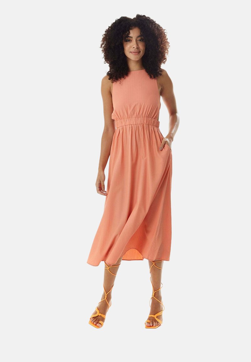 Peach-colored, sleeveless midi dress with a cinched waist, side pockets, and a flowing skirt. Paired with orange strappy sandals.