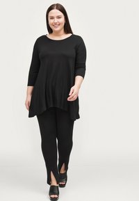 Sort tunika med trekvartærmer og asymmetrisk kant, iført sammen med sorte leggings med sideslids, båret med sorte hælede sandaler.