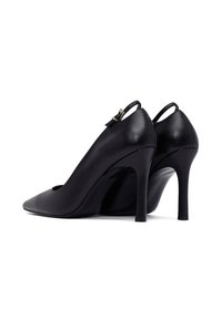 Zapatos de tacón alto de charol negro con puntera puntiaguda y pequeñas correas con hebillas doradas sobre un fondo blanco.