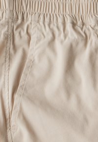 Tissu beige avec une texture lisse, présentant une taille élastique et des coutures le long des coutures, caractéristiques d'un design de shorts décontractés.