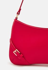 Červená kožená kabelka přes rameno se zlatým detailem na popruhu na bílém pozadí.