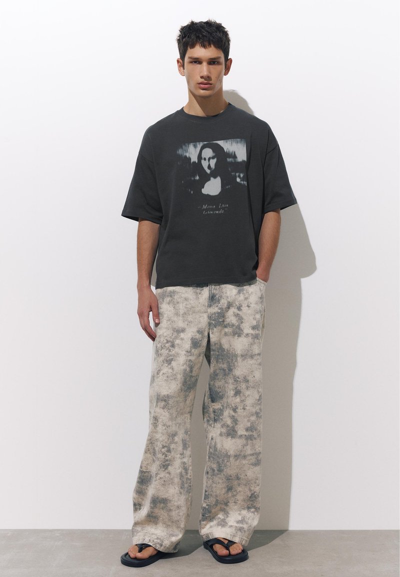 Jeune homme portant un T-shirt noir oversize avec une impression Mona Lisa et un pantalon large tie-dye, debout contre un mur blanc uni.