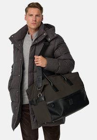 Sort og mørkebrun duffelbag med læderdetaljer, skulderrem og fremtrædende brandlogo. Materialet virker holdbart og struktureret.
