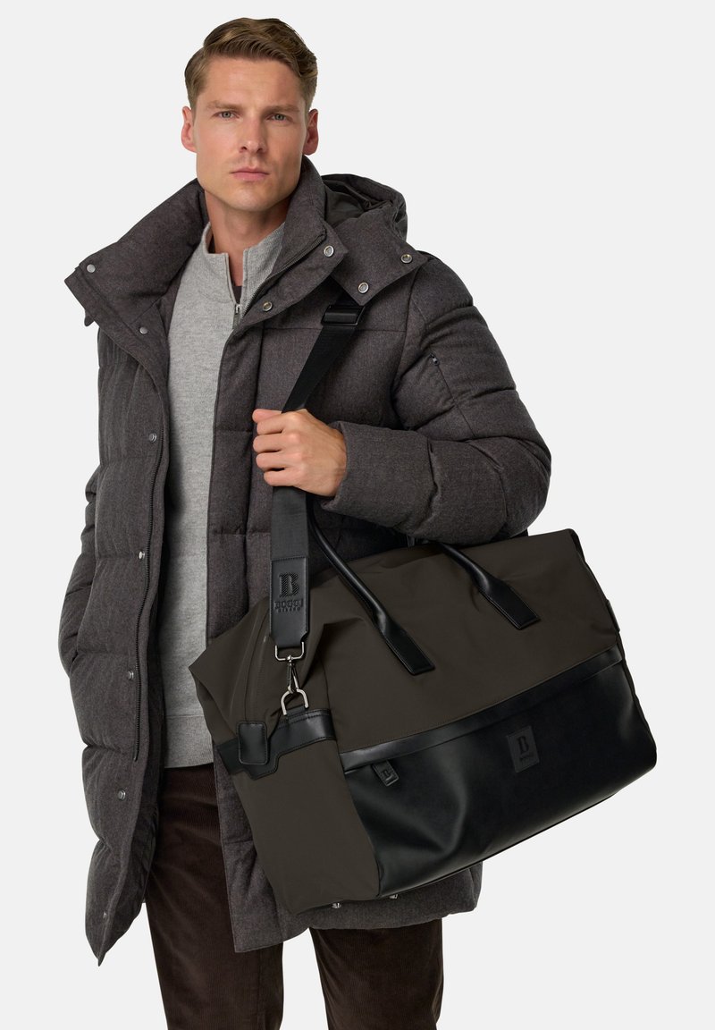Sort og mørkebrun duffelbag med læderdetaljer, skulderrem og fremtrædende brandlogo. Materialet virker holdbart og struktureret.