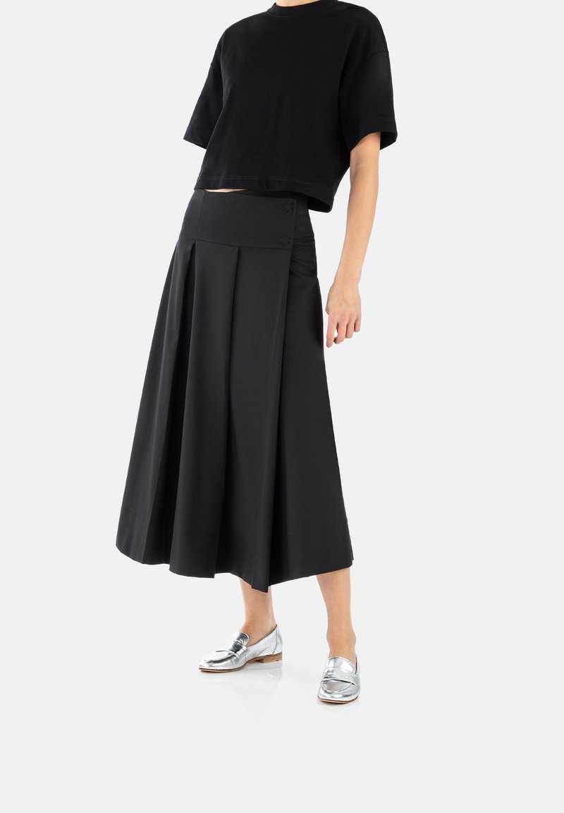 Femme portant un top court noir à manches courtes, une jupe midi plissée noire et des mocassins argentés sur fond blanc.