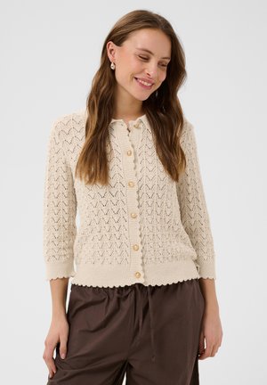 Femme souriante, portant un cardigan boutonné en maille crème à bord festonné et un pantalon marron à cordon, debout devant un fond uni.