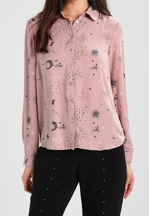 Femme portant une chemise rose à manches longues boutonnée avec des motifs d'étoiles et de constellations en noir et blanc et un pantalon noir avec de petits points blancs.