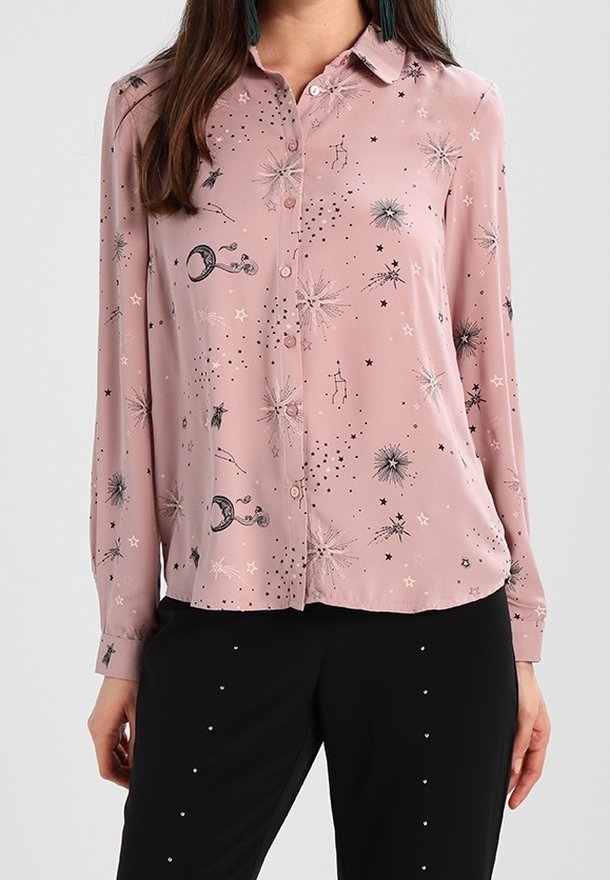 Femme portant une chemise rose à manches longues boutonnée avec des motifs d'étoiles et de constellations en noir et blanc et un pantalon noir avec de petits points blancs.