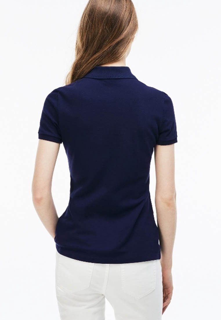 Femme aux cheveux longs portant un polo bleu marine à manches courtes ajusté et un pantalon blanc, vue de dos sur un fond uni.