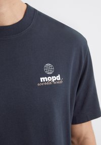 Marine bomull t-skjorte med korte ermer, med et hvitt globusgrafikk og teksten "mopd world.wide" i lys beige. Glatt tekstur.