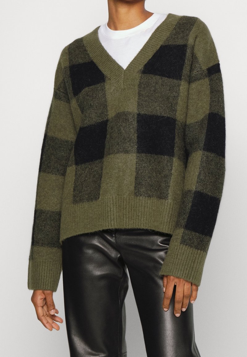 Personne portant un pull à col en V à carreaux vert olive et noir par-dessus une chemise blanche, associé à un pantalon en cuir noir.