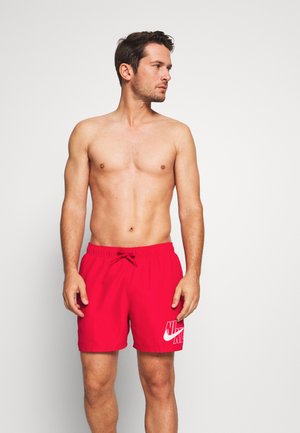 Oberkörperfreier Mann mit kurzen dunklen Haaren, der rote Nike-Badeshorts trägt, steht vor weißem Hintergrund und blickt nach links.