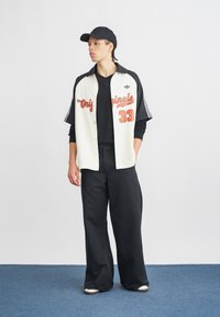 Maglietta da baseball in crema e nero con scritte arancioni, abbinata a pantaloni neri larghi. Gli accessori includono un berretto nero.