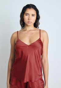 Camisole en satin rouge avec des bretelles spaghetti réglables, présentant un décolleté en V et une silhouette fluide. Texture lisse avec un éclat subtil.