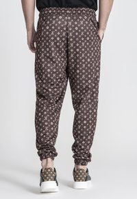 Pantalones marrones con un diseño de estampado repetido del logo, cintura elástica y puños ajustados. Combinados con zapatillas negras y beige.
