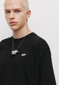 Sort bomulds T-shirt med korte ærmer, der har et lille hvidt logo på brystet. Modellen bærer et sølvfarvet, flerlags halskæde.