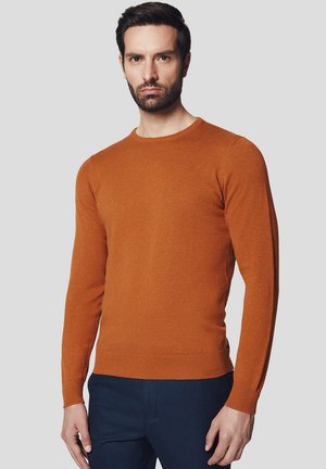 Sweter w kolorze rdzy, wykonany z miękkiej dzianiny, z okrągłym dekoltem i długimi rękawami, z prążkowanymi mankietami i dołem. Noszony z granatowymi spodniami.