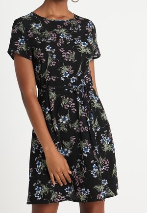 Femme portant une robe noire à manches courtes avec un motif floral bleu et rose et une ceinture à la taille, debout avec une main reposant sur la jupe.
