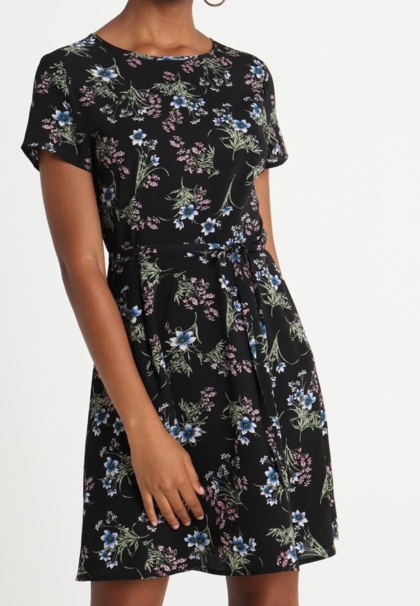 Femme portant une robe noire à manches courtes avec un motif floral bleu et rose et une ceinture à la taille, debout avec une main reposant sur la jupe.