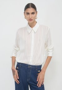 Donna con capelli slicked-back, che indossa una blusa bianca trasparente a righe, jeans blu a vita alta con bottoni laterali e orecchini a cerchio dorati.