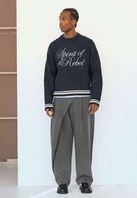 Trui in donker marineblauw met witte letters 'Spirit of a Rebel', gecombineerd met grijze plissébrogues en zwarte schoenen. Duidelijke gladde stoffen texturen.