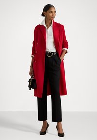 Palton roșu cu două rânduri de nasturi peste o cămașă albă, asortat cu pantaloni negri adaptați și tocuri negre, ținând o geantă mică neagră.