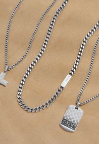 Tres collares de plata: uno con una cruz, uno de cadena gruesa y uno con una etiqueta tipo perro texturizada. Todos presentan diferentes estilos de eslabones y acabados.