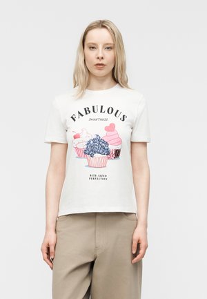 Giovane donna che indossa una T-shirt bianca con illustrazioni colorate di cupcake e la scritta "Dolcezza favolosa, perfezione in bocconcini", pantaloni beige.
