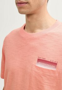 Kortærmet, lyserød t-shirt i et blødt stof. Har en lomme med et trykt design. Ribbet halsudskæring tilføjer tekstur.