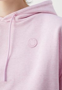 Gros plan d'une personne portant un sweat à capuche rose clair avec un petit emblème de visage souriant brodé sur la poitrine et des cordons suspendus.