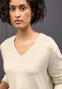 Krémově zbarvený, měkký pletený svetr s výstřihem do V a tříčtvrtečními rukávy, který se vyznačuje hladkou texturou a minimalistickým designem.
