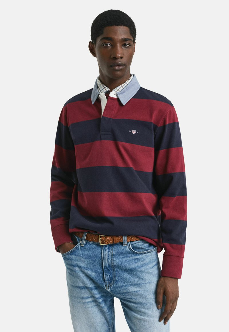 Junger Mann in einem rot-blauen gestreiften Rugbyshirt mit einem hellblauen Kragen über einem karierten Hemd, kombiniert mit blauen Jeans und einem geflochtenen braunen Gürtel.