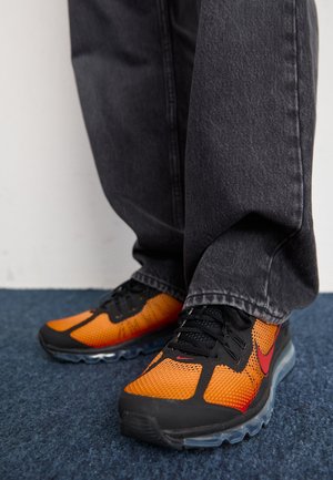 Zapatillas deportivas negras y naranjas con parte superior de malla, cordones negros y suela transparente, usadas con vaqueros grises holgados sobre una superficie alfombrada azul.