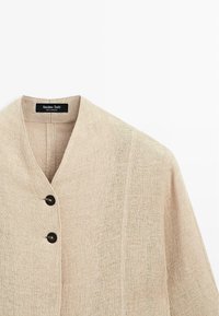 Beige linnedragt med sjalskrave, der har to sorte knapper og en ren finish. Minimalistisk design med subtil tekstur.