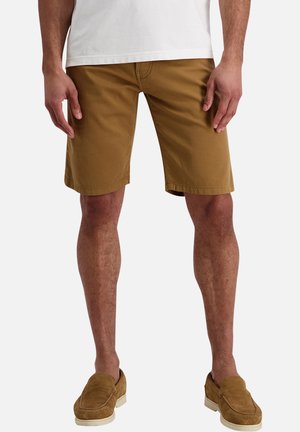 Mandlig model iført brune knælange shorts, hvid t-shirt og matchende brune slip-on sko mod en ensfarvet baggrund.