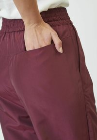Bordo hlače s elastičnom trakom u struku i bočnim džepovima, izrađene od glatkog, laganog materijala s opuštenim krojem.