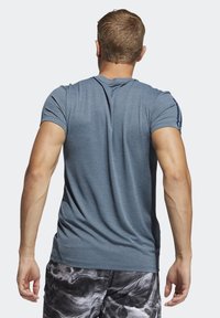 Hombre con una camiseta de textura azul y pantalones cortos de patrón negro y gris, de pie de espaldas contra un fondo claro liso.