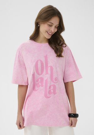 Fiatal nő mosolyogva lefelé néz, laza rózsaszín "oh la la" feliratú pólót és fehér nadrágot visel, kezén fekete karpereccel.