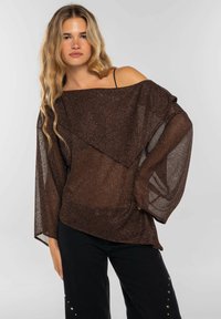 Top marrón de punto metálico con diseño de hombros descubiertos, mangas amplias y aspecto en capas. Combinado con pantalones negros que presentan tachuelas plateadas como acento.