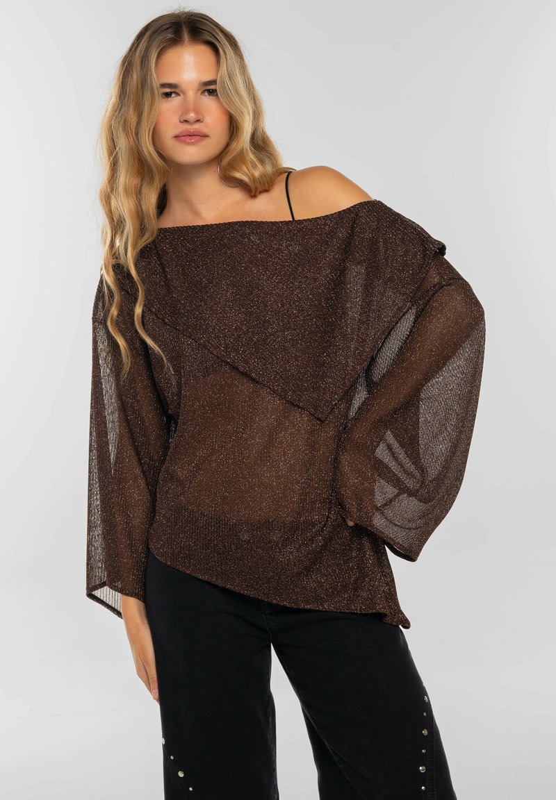 Top marrón de punto metálico con diseño de hombros descubiertos, mangas amplias y aspecto en capas. Combinado con pantalones negros que presentan tachuelas plateadas como acento.