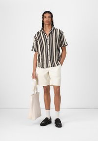 Camisa de mangas cortas a rayas en blanco y negro, pantalones cortos color crema, mocasines negros, calcetines blancos, sosteniendo una bolsa tote beige, detalles de tela texturizada.