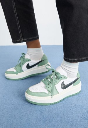 Person iført hvide og mintgrønne Nike sneakers med hvide sokker og sorte korte bukser, stående på et blåt tæppe.