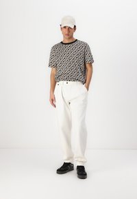 Μαύρη και λευκή καρώ κοντομάνικη μπλούζα, φαρδιά λευκά παντελόνια, μαύρα αθλητικά παπούτσια και καπέλο σε ανοιχτό χρώμα.