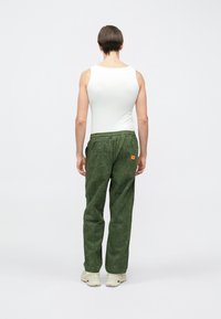 Pantalones de pana verdes con una cintura elástica, que cuentan con dos bolsillos traseros y un parche de logo naranja; combinados con una camiseta sin mangas blanca ajustada.