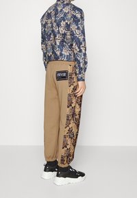 Blommigt mönstrad blå skjorta med långa ärmar i kombination med beiga sweatpants som har en blommig sidodesign och en etikett på baksidan, bärs med svarta sneakers.