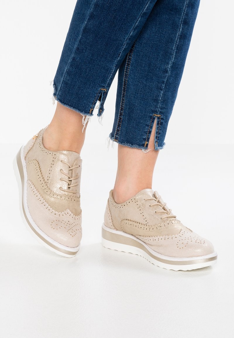 Baskets beiges de style brogue avec semelles compensées blanches portées avec un jean bleu raccourci, muni d’ourlets effilochés et de fentes à l’avant des chevilles.