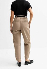 Pantalon beige taille haute avec revers, coupe ajustée et poches arrière. Porté avec des chaussures noires et un haut noir simple.