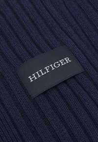 Tessuto a coste navy con una etichetta rettangolare nera che presenta "HILFIGER" in lettere bianche, sfondo testurizzato e scritta leggermente in rilievo.