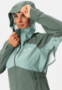 Giacca da trekking impermeabile in verde bicolore, con cappuccio strutturato, zip frontale e zip di ventilazione. Tessuto liscio e leggero.
